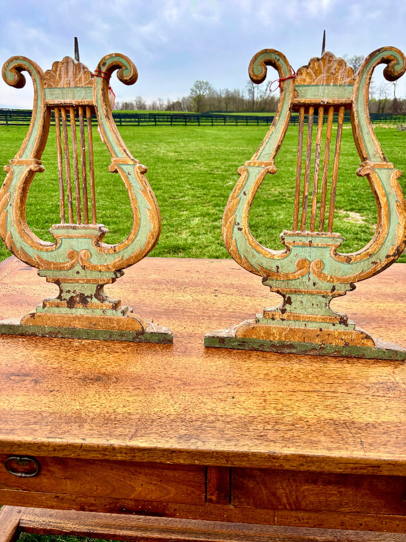 Two Antique Italian ornate lyre-shaped candlesticks on a wooden surface with a grassy field and trees in the background.