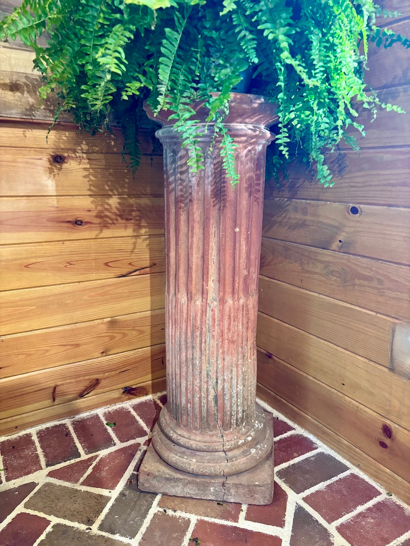 Decorative terra cotta column with a potted fern against a wooden fence.